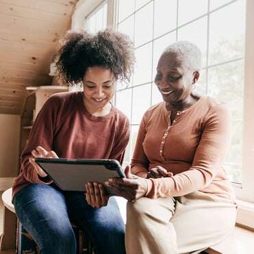 Daughter and senior mother sit by window smiling and looking at tablet