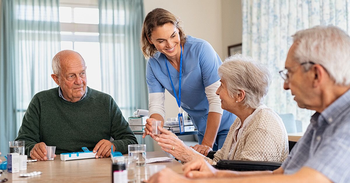 Nurse in scrubs hands senior woman medicine in cup at dining room table