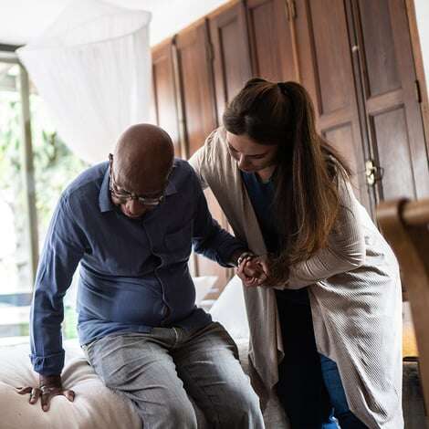 Woman aid helps senior man sit down on edge of bed at home