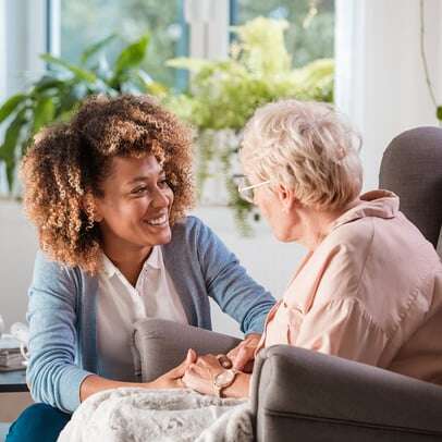 Young woman crouches next to elderly woman in chair as they hold hands