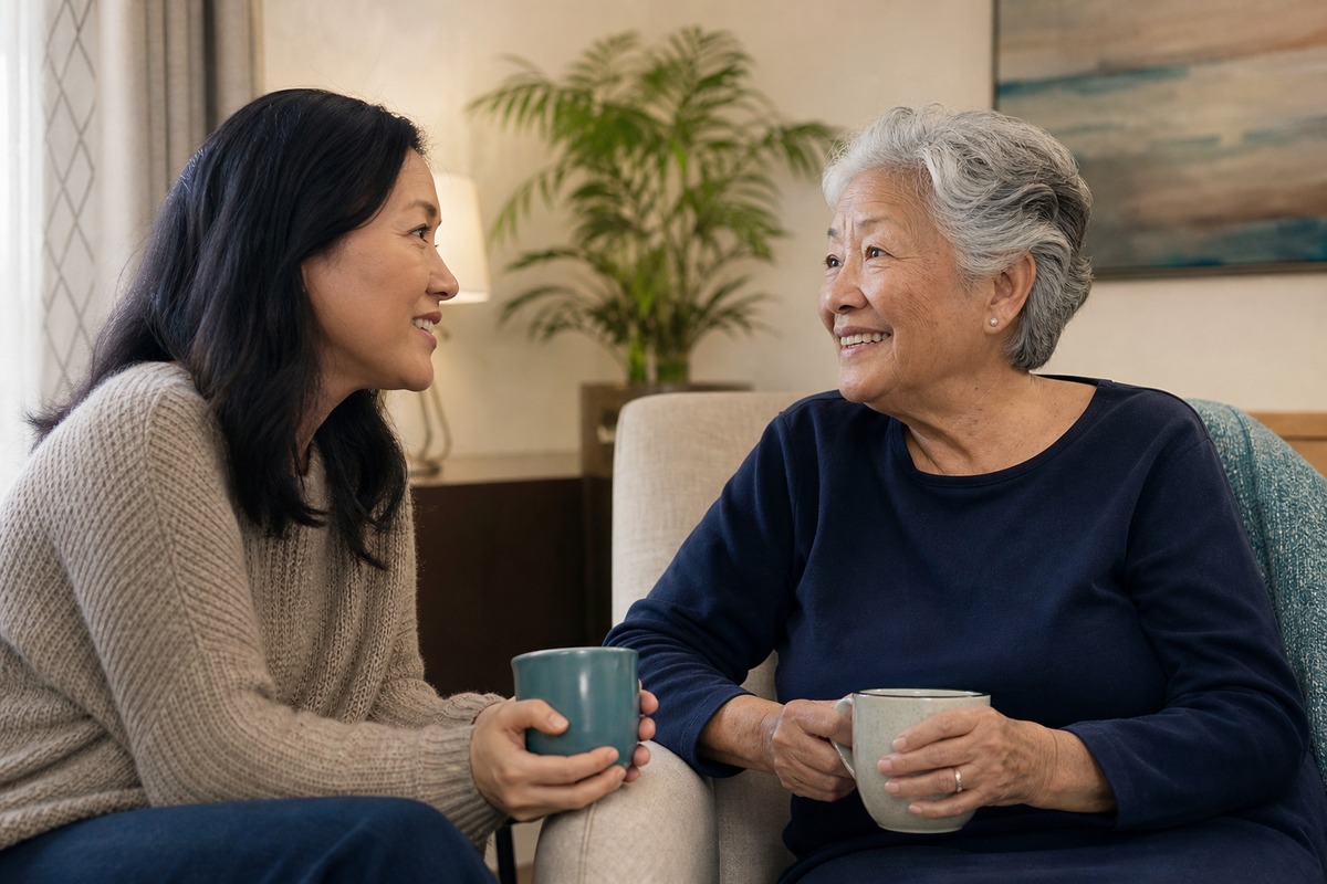 An adult child visits their older parent in assisted living, smiling and enjoying tea together.